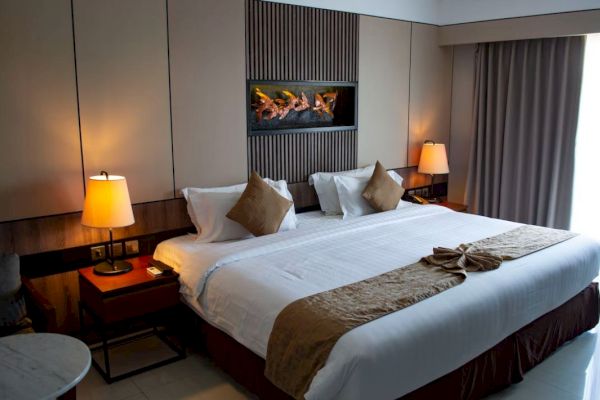 A neatly arranged hotel room features a large bed with white and beige linens, two bedside tables with lamps, and a piece of wall art.