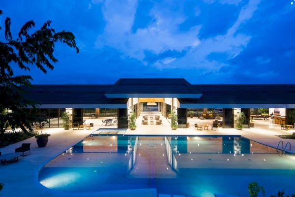 A modern, illuminated building with a wide swimming pool in front, surrounded by lounge areas and greenery, captured during twilight hours.