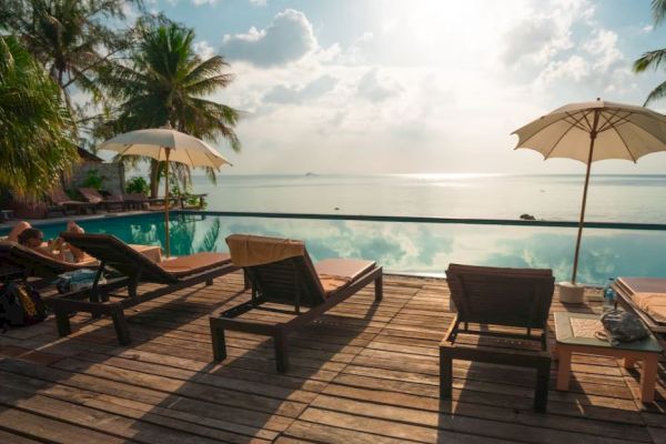 A serene tropical poolside scene with lounge chairs, umbrellas, palm trees, and a stunning view of the ocean under a partly cloudy sky.