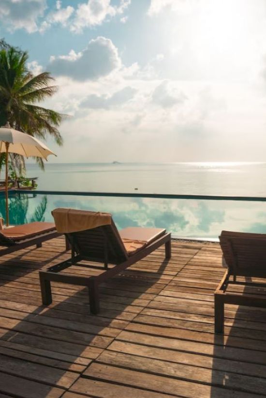 A wooden deck by an oceanside pool, with lounge chairs, umbrellas, palm trees, and a serene view of the sky and water under the sun.