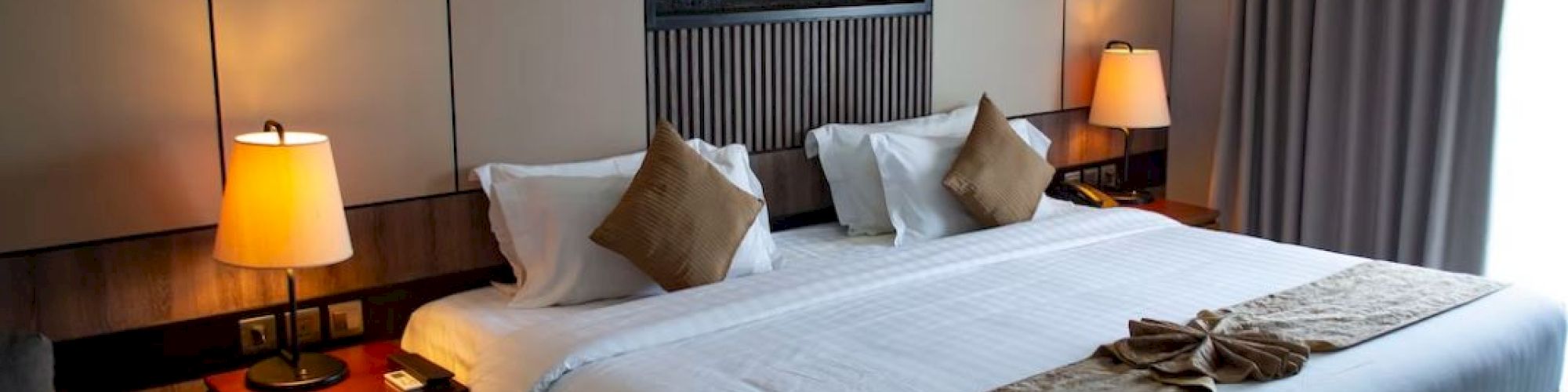 A cozy hotel room with a large bed, two lit bedside lamps, pillows, a wooden headboard, and a window with closed curtains, creating a warm ambiance.