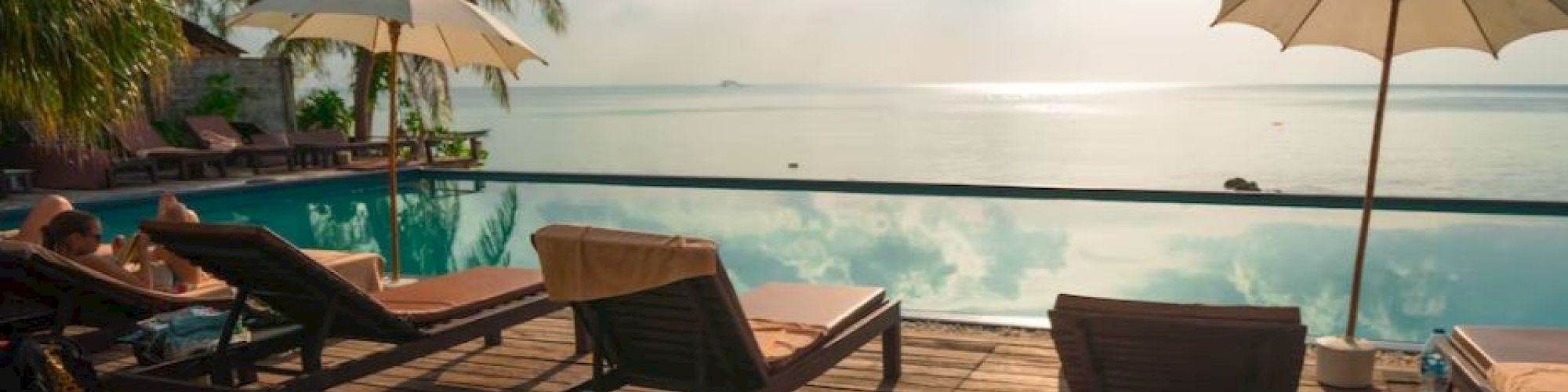 A serene poolside scene with lounge chairs, umbrellas, and palm trees, facing a tranquil ocean under a slightly cloudy sky with bright sunlight.