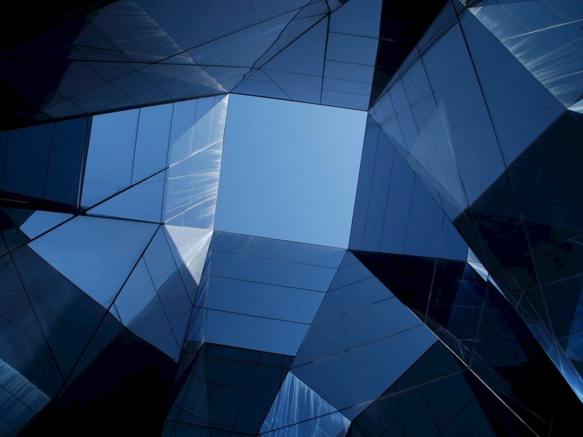 The image features a view looking upwards at a modern building with reflective, angular glass panels, revealing a clear blue sky in the center.