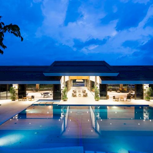 A luxurious building with an illuminated outdoor pool is shown under a twilight sky, reflecting its lights on the water.