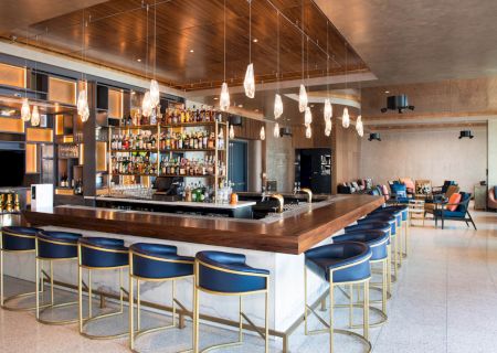 A modern bar with a central counter, numerous liquor bottles, stylish seating, pendant lights, and a lounge area with couches and tables.