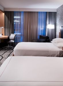 The image shows a modern hotel room with two beds, a wall-mounted TV, a desk, chair, lamps, and patterned carpet flooring.