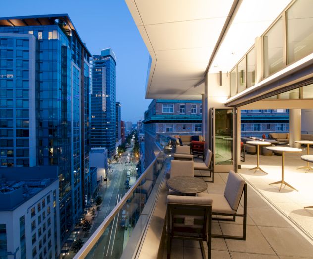 The image shows a modern cityscape with tall buildings at dusk. There is a spacious balcony with tables and chairs overlooking the street below.