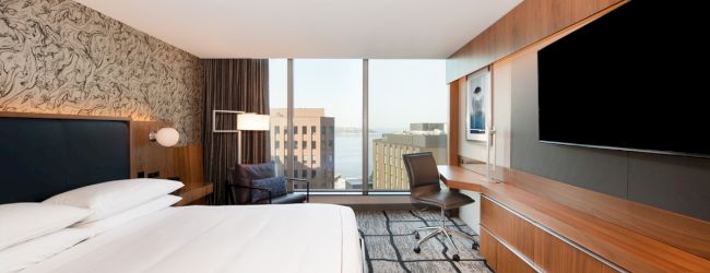 A modern hotel room with a large bed, a desk, a flat-screen TV, and a window offering a city view and some water beyond the buildings.