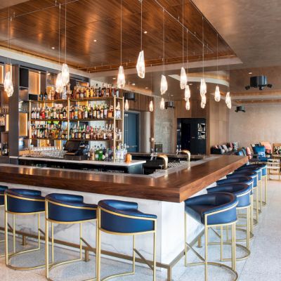 This image shows a modern bar with stylish seating, a well-stocked bar, hanging lights, and a cozy seating area in the background.