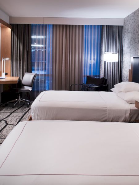 A modern hotel room with two neatly made beds, a wall-mounted TV, a desk with a chair, and patterned wallpaper, with a window featuring closed curtains.