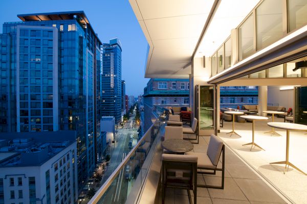 The image shows a modern cityscape with tall buildings and a balcony with tables and chairs, overlooking a street at dusk or early evening.