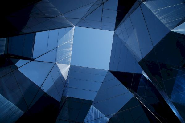 The image shows a view looking up through a geometric arrangement of reflective glass panels, revealing a patch of clear blue sky.