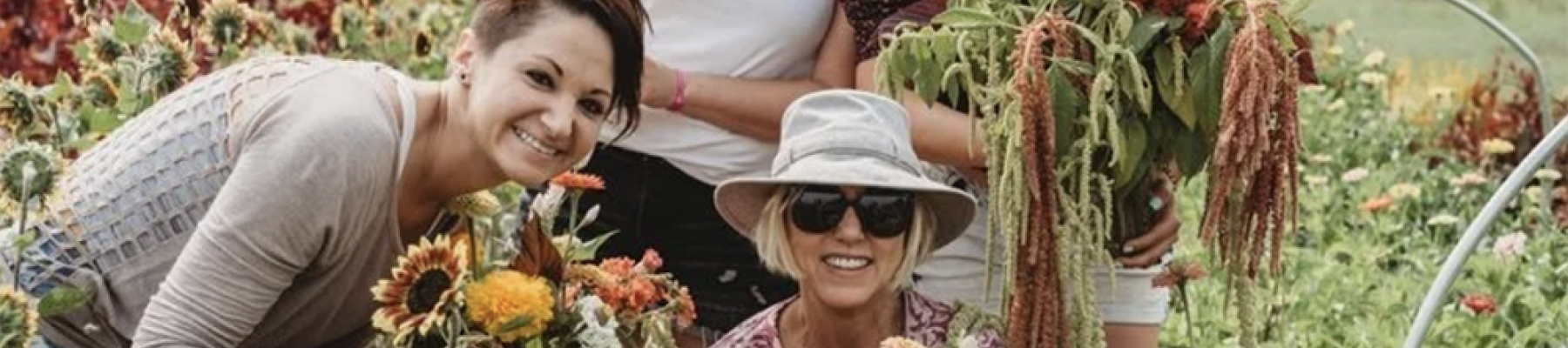 A group of four women is in a garden, holding bouquets of freshly picked flowers and smiling at the camera.