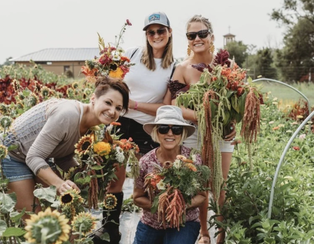 A group of four women is in a garden, holding bouquets of freshly picked flowers and smiling at the camera.