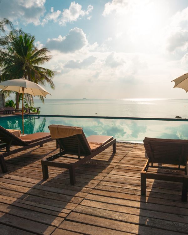 A serene beach view with lounge chairs, umbrellas, a pool, and palm trees under a sunny sky. The ocean is visible in the background.