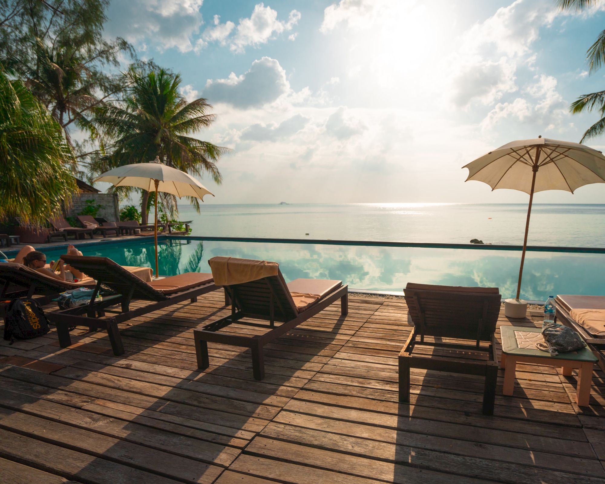 Lounge chairs on a wooden deck face a serene pool and beachfront, with palm trees and umbrellas under a partly cloudy sky at sunset.