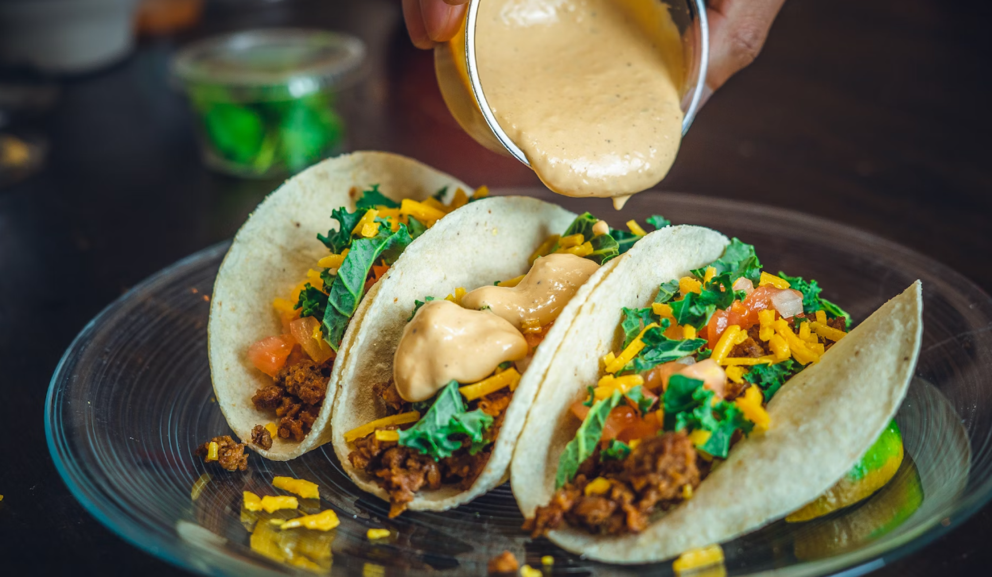 The image shows three tacos on a glass plate, being topped with creamy sauce. The tacos are filled with vegetables, meat, and shredded cheese.
