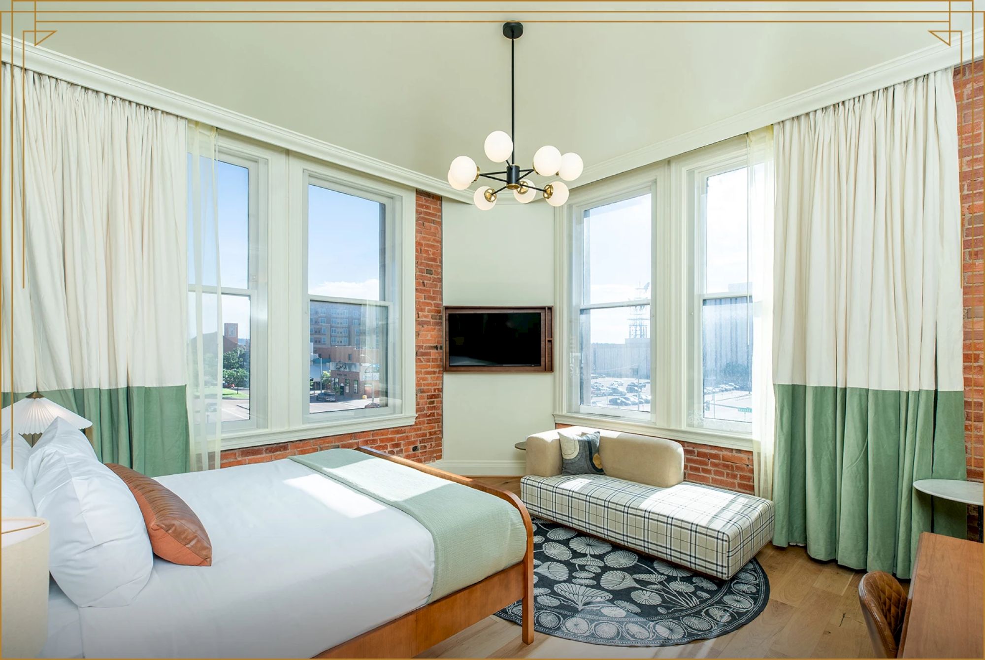 A bright, modern room with large windows, a cozy bed, a TV, and a seating area featuring a small sofa and table, illuminated by a chic chandelier.