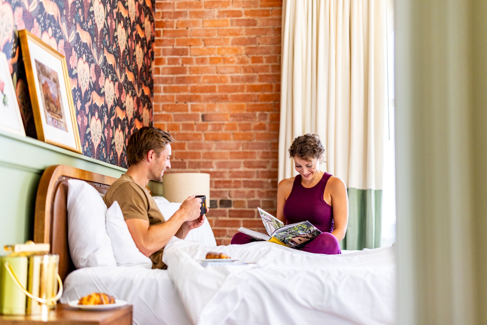 Two people are sitting on a bed in a cozy bedroom. One is holding a magazine while the other has a mug. Both seem relaxed and happy.
