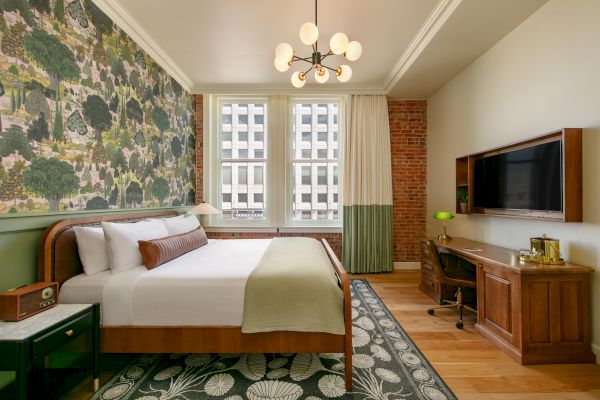 A stylish bedroom with a large bed, patterned feature wall, desk with a TV, and a city view through two windows, blending modern and vintage elements.