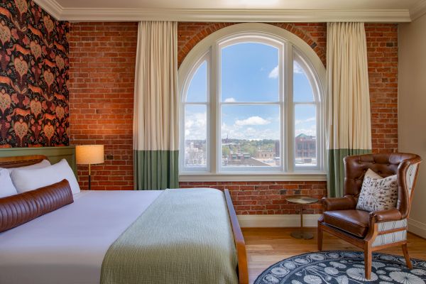 A cozy room with a bed, leather chair, and large arched window with city view. There's exposed brick, wallpaper, and patterned rug.
