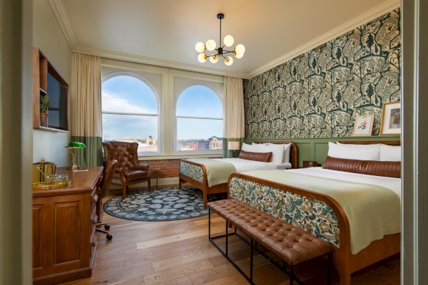 A bedroom with two beds, a desk, a leather chair, a patterned rug, and large windows with a city view. The room features decorative wallpaper.