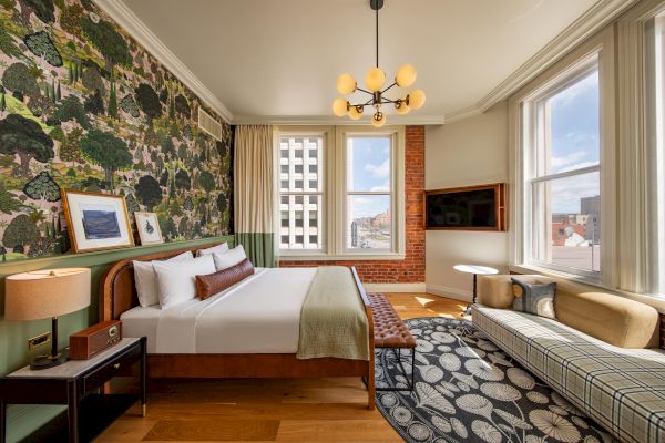 This image shows a stylish bedroom with a large bed, patterned wallpaper, a modern chandelier, large windows, a TV, and a lounge area.