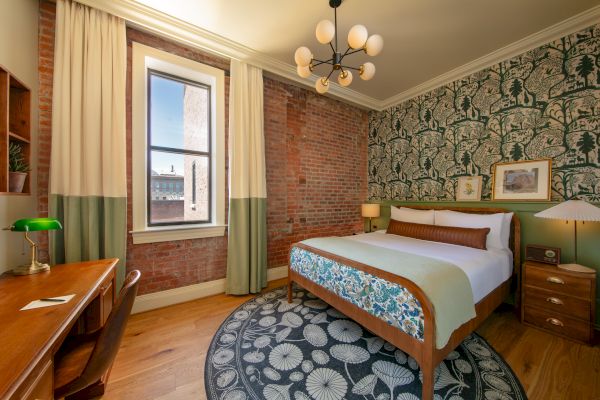 A bedroom with a bed, a green and white decorative theme, brick and wallpapered walls, a desk, a window with curtains, and a round rug on the floor.