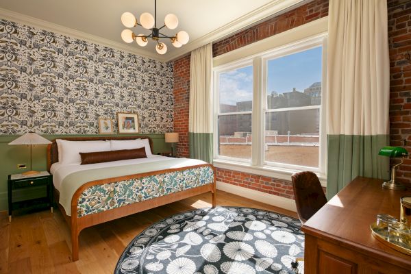 A cozy bedroom with a bed, decorative wallpaper, a round rug, a wooden desk, and large windows letting in natural light, ends the sentence.