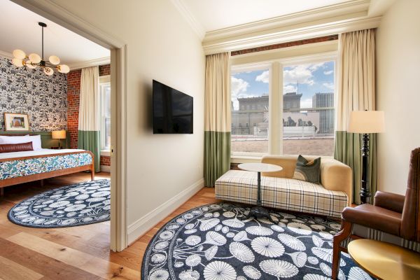 The image shows a modern hotel room with a seating area, large windows, patterned rugs, and a flat-screen TV. There is a view of a building outside.