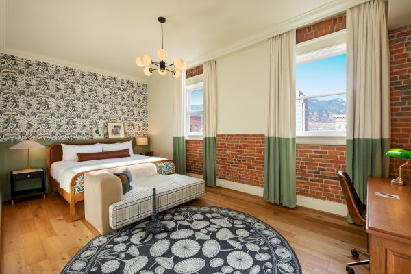 A stylish bedroom with a double bed, patterned wallpaper, brick walls, large windows, modern lighting, a desk, and a cozy bench.
