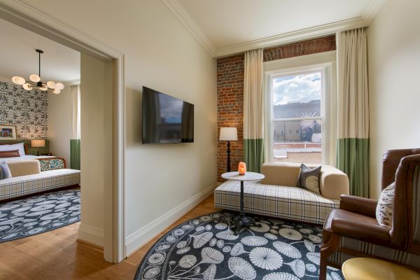 The image shows a cozy living area with a wall-mounted TV, a round table, and a sofa, leading to a decorated bedroom with a patterned rug and wallpaper.