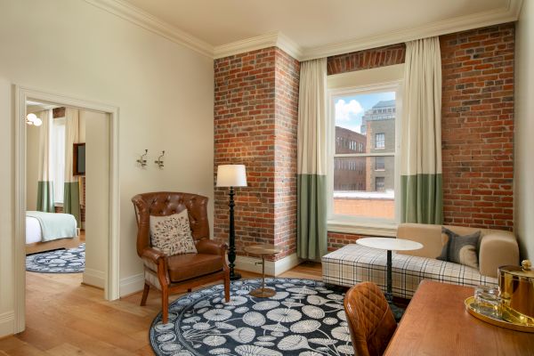 A cozy living space with a brick accent wall, window, armchair, sofa, area rug, and adjacent bedroom visible through an open door.