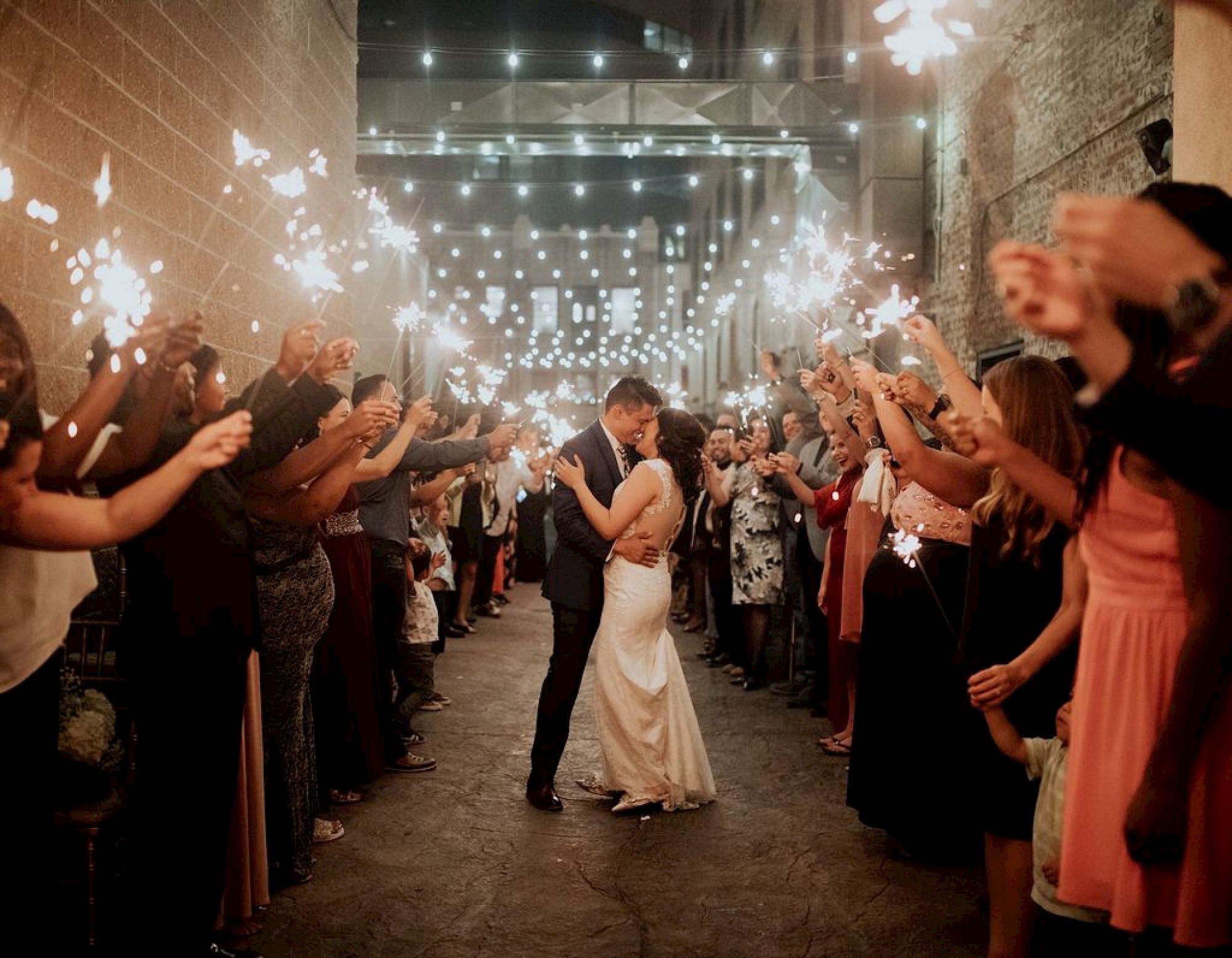 A bride and groom kiss in the middle of an aisle as guests hold sparklers around them, with string lights hanging above.