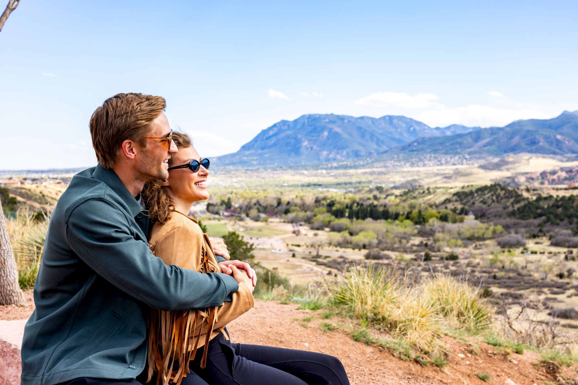 A couple sits together on a hilltop, smiling and enjoying a scenic view of a sprawling valley with mountains in the background on a clear day.
