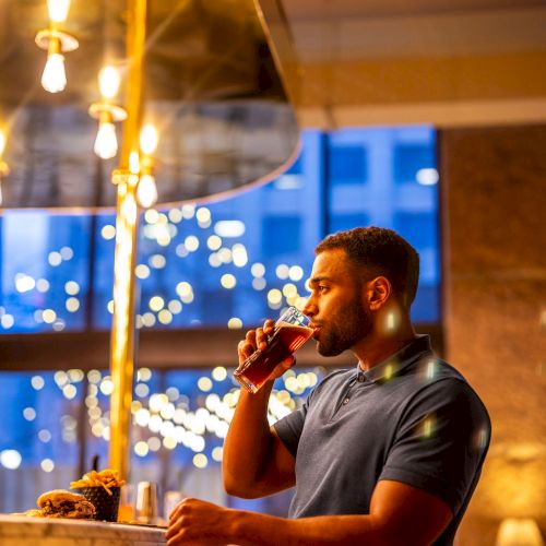 A man sits at a bar, drinking a beverage with decorative lights and a large window in the background. He appears relaxed and thoughtful.