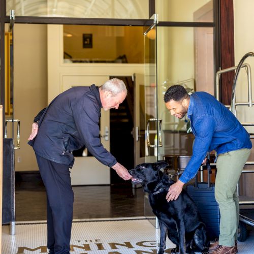Two men stand at an entryway, one bending to pet a black dog while the other holds the dog on a leash, with clear glass doors behind them.