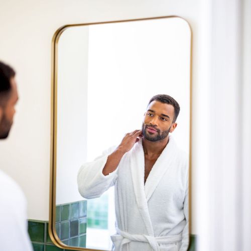A man in a white robe looks at himself in a mirror, adjusting his collar in a bathroom with green tiles and a gold-framed mirror on the wall.