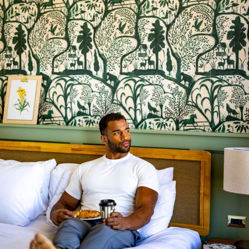 A person sits on a bed, holding a mug and a plate, against a backdrop of green patterned wallpaper in a cozy room.