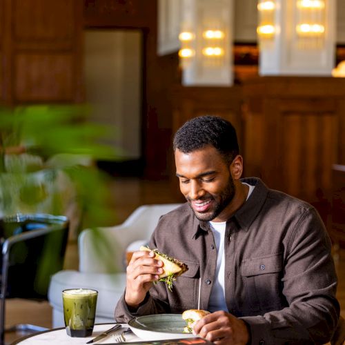 A person is sitting at a table in a cozy room, eating a sandwich with a drink nearby. A book titled 