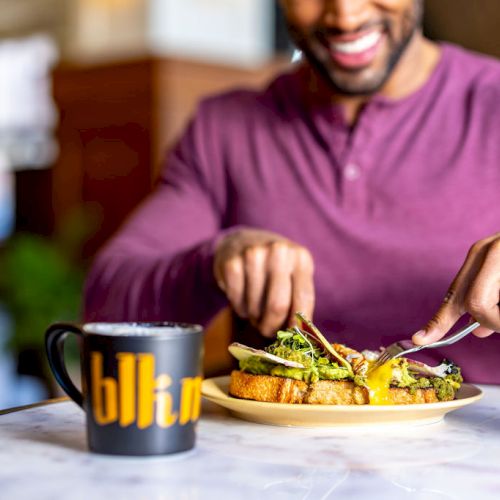 A person in a maroon shirt is cutting food on a plate with a fork and knife, with a mug labeled 