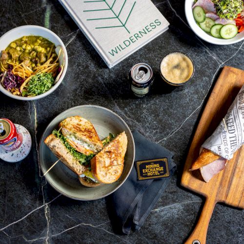 The image shows sandwiches, a salad, a can of beer, a latte, and a book titled 