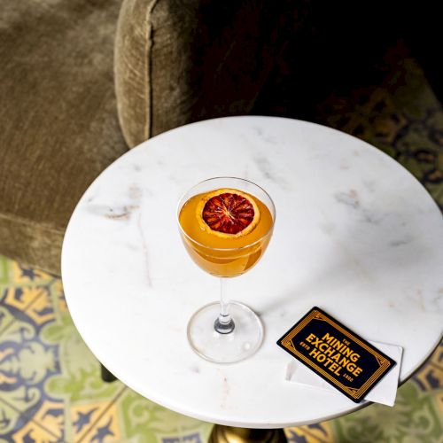 A round marble table with a cocktail featuring a blood orange slice, a card with text, and a comfortable chair in the background.