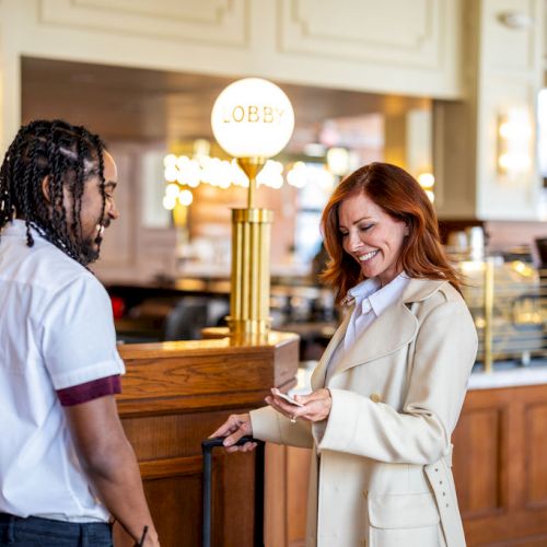 A woman in a white coat appears to be checking in at a hotel or restaurant, greeted by a staff member wearing a uniform with a name tag.