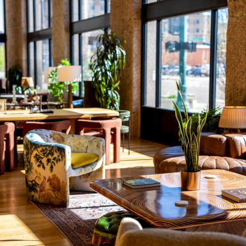 A brightly lit lounge area with large windows, wooden tables, comfortable seating, plants, and books on tables, creating a cozy ambiance.