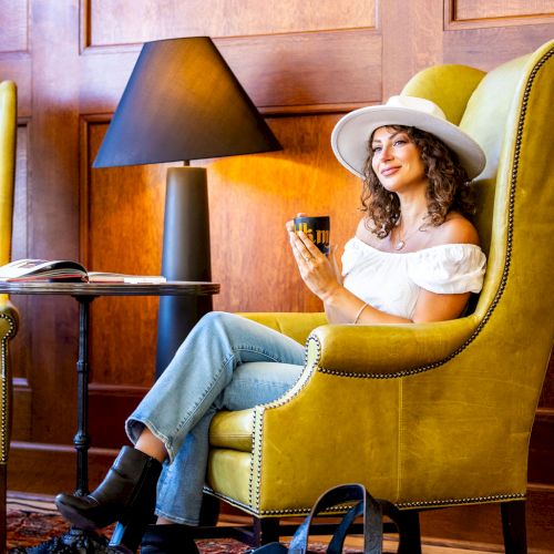 A woman in a hat sits on a yellow chair, holding a cup, with a black bag on the floor beside her and a lamp on the side table nearby.
