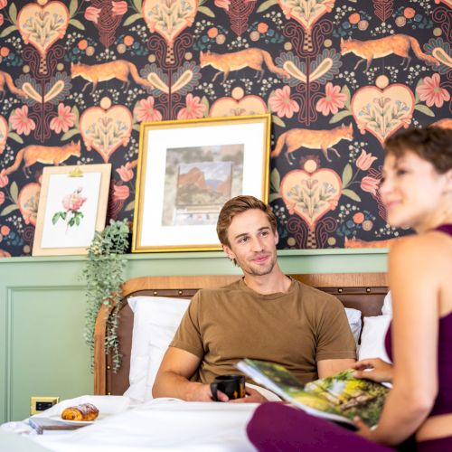 A man and woman are sitting on a bed with floral wallpaper and framed art in the background, engaging in a relaxed conversation or activity.