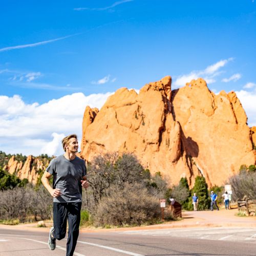 A person is jogging on a road with stunning red rock formations and a bright blue sky in the background. Other people can be seen in the distance.