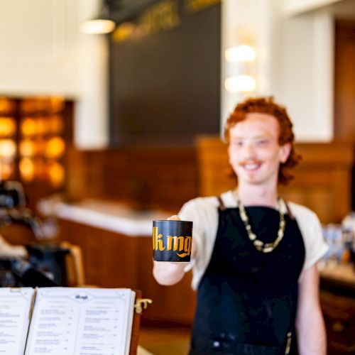 A person standing behind a counter in a well-lit café, holding out a mug, with a menu visible on the left side of the image.