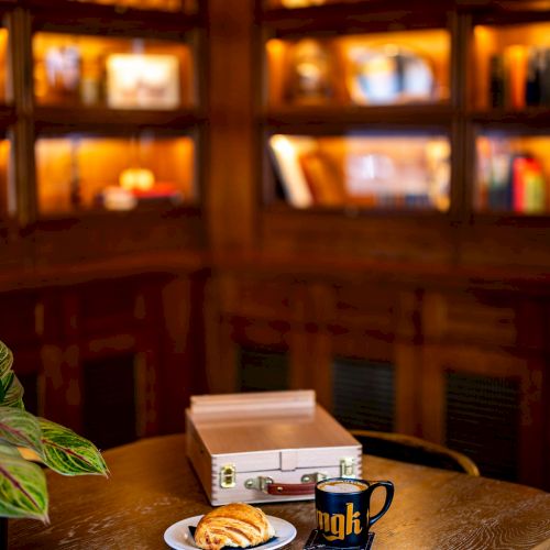 A cozy room with dim lighting, featuring a table with a plate of pastries, a mug, and an open briefcase, and a bookshelf in the background.
